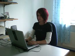 Frau mit rotem Haar arbeitet am Laptop in einem Büro mit blauem Vorhang.