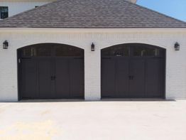 Double garage doors with dark wood finish on a white brick building, under a shingled roof.