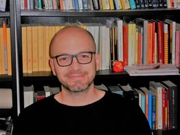 Uomo con occhiali davanti a una libreria piena di libri colorati.