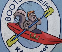 Karikatur eines Eichhörnchens im roten Kanu mit gelbem Paddel, umgeben von "Bootsverleih Königsboot".