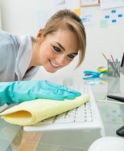 Frau reinigt Tastatur am Schreibtisch mit gelbem Tuch, trägt blaue Handschuhe und lächelt.