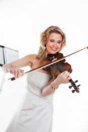 Frau in weißem Kleid spielt Geige, lächelt, mit Notenständer im Hintergrund.