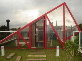 Estrutura triangular de vidro e metal vermelho em jardim, com céu nublado ao fundo.
