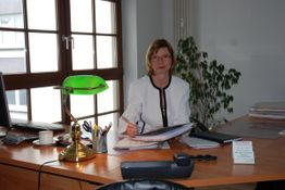 Frau in weißem Blazer sitzt am Schreibtisch mit grünem Lampenschirm, Akten und Computer.
