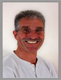 Ein lächelnder Mann mit Brille und Schnurrbart vor hellem Hintergrund.