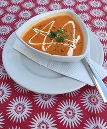 Sopa de tomate en plato triangular sobre mantel rojo con diseño floral, decorada con crema y hierbas.