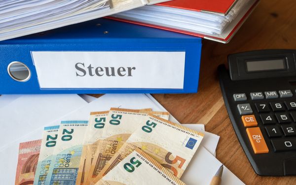 Ordner mit der Aufschrift „Steuer“, Euro-Banknoten und Taschenrechner auf einem Schreibtisch.