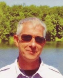 Ein Mann mit Sonnenbrille steht vor einem See und Bäumen im Hintergrund.