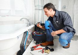 Plumber fixing bathtub pipes, surrounded by tools in a bright bathroom.