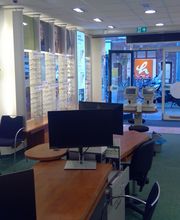 Optiekwinkelinterieur met stoelen, display met brillen en een bureau met computers.