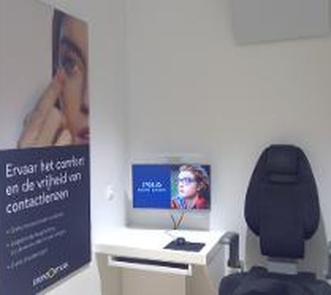Kamer met een oogmeetstoel, poster over contactlenzen en een computermonitor met een gezicht.