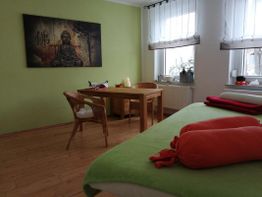 Meditationsraum mit Buddha-Bild, grünem Bett, Tisch und Korbstühlen, helles Fensterlicht.