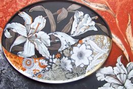 Ovaler Teller mit weißen Blumenmotiven auf schwarzem, orangefarbenem und rotem Hintergrund.