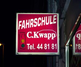 Fahrschulschild mit der Aufschrift "C. Kwapp", Telefonnummer und Ampelgrafik bei Nacht.