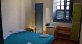 Quarto de hotel simples com cama de casal, espelho, cabideiro amarelo e janela.