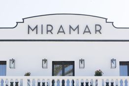 Fachada blanca de un edificio con el letrero "Miramar" y balcones con plantas.