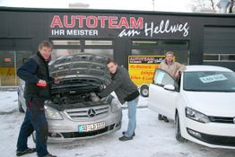 Eine Autowerkstatt mit zwei Männern, die den Motor eines silbernen Mercedes inspizieren.