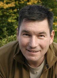 Ein Mann lächelt in die Kamera, im Hintergrund sind grüne Blätter und Herbstfarben zu sehen.