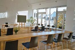 Heller Seminarraum mit Holztischen und Stühlen, großer Fensterfront und grünem Flipchart.