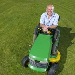Man smiling while sitting on a green riding lawn mower on a sunny day.