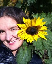 Eine Frau lächelt neben einer großen, gelben Sonnenblume im Freien.