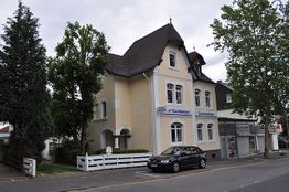 Gelbes Gebäude mit Fachwerkgiebel, Auto auf der Straße, Bäume und Wolken im Hintergrund.