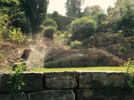Terrassierter Garten mit grünen Büschen und Steinmauern, im Sonnenlicht mit Sprinklerbewässerung.
