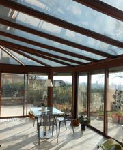 Wintergarten mit Glasdach, Tisch und Stühlen, umgeben von Pflanzen und großer Fensterfront.