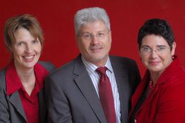 Drei Personen in Anzügen vor rotem Hintergrund, lächeln in die Kamera.