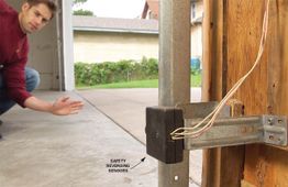 Man crouching in garage examines safety reversing sensors on a door frame.