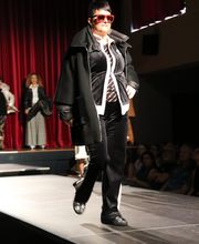 Person auf Laufsteg in schwarzer Kleidung und roter Brille, roter Vorhang im Hintergrund.