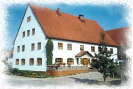 Großes weißes Haus mit rotem Dach und vielen Fenstern, umgeben von Gartenpflanzen.