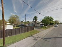 Residential street with houses, a fence, palm tree, and power lines under a cloudy sky.
