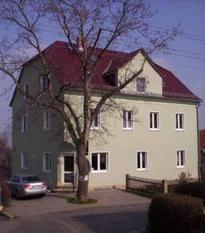 Eckhaus mit rotem Dach, grünem Putz und kahlem Baum davor; daneben parkt ein silbernes Auto.