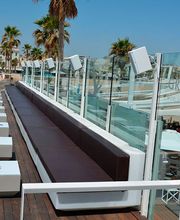 Terraza de playa con sofás marrones, mesas blancas y palmeras al fondo. Ambiente soleado.