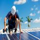 Worker in a hard hat installing solar panels on a roof under a sunny blue sky.
