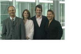 Vier Personen in Businesskleidung lächeln in die Kamera vor einem modernen Bürogebäude.