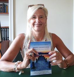 Una donna sorride, seduta a un tavolo, e tiene un libro intitolato "Ipnosi e Mindfulness".