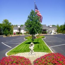 Parking lot with a flagpole, statue, and flowers leading to a building under a clear blue sky.