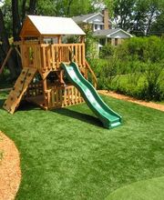 Holzspielplatz mit Schaukel, Kletterwand und Rutsche auf grünem Rasen, umgeben von Bäumen.
