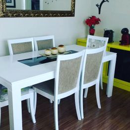 Sala de jantar com mesa branca, cadeiras estofadas, flores vermelhas e espelho na parede.