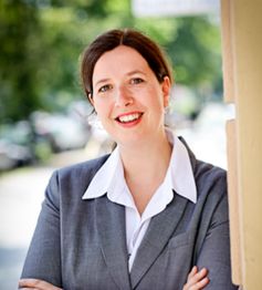 Frau in grauem Anzug lächelt freundlich, steht draußen vor einer unscharfen, grünen Kulisse.