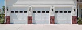 Three white garage doors with brick accents beneath exterior lamps.