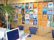 Reisebüro mit Broschüren an der Wand, Computer auf dem Tisch und Topfpflanze daneben.