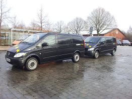 Zwei schwarze Vans mit Stern-Logo auf nassem Pflaster vor kahlen Bäumen geparkt.