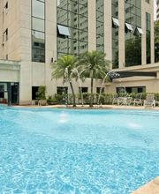 Piscina em frente a edifício moderno, com cadeiras ao redor e área verde ao fundo.