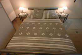 Graues Doppelbett mit gemustertem Bettbezug, zwei Nachttische mit Lampen, gefliester Boden.