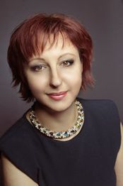 Eine Frau mit kurzem, rötlichem Haar und schwarzem Oberteil vor grauem Hintergrund.