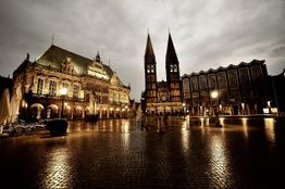 Abendansicht eines beleuchteten Rathauses und Doms an einem regennassen Platz in Bremen.