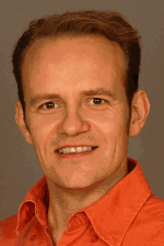 Porträt eines lächelnden Mannes in einem orangefarbenen Hemd vor einem neutralen Hintergrund.